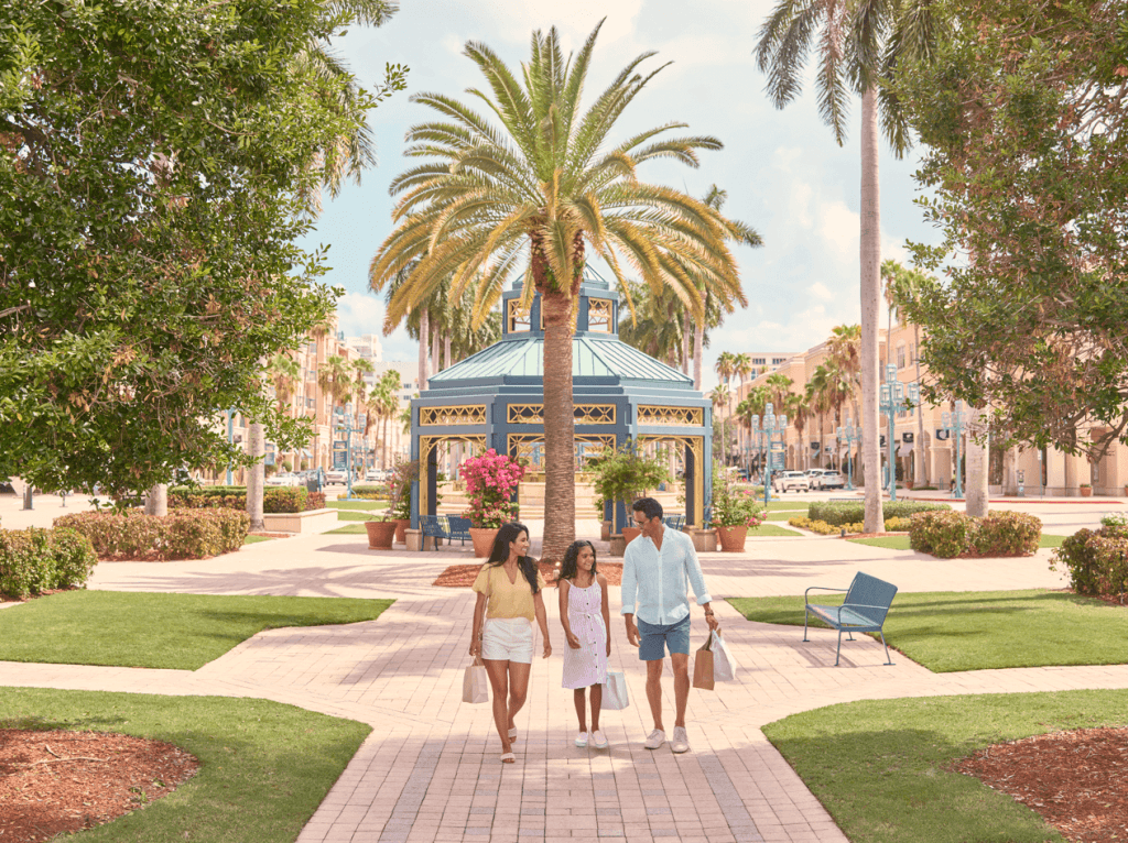 A family of three strolls along a sunlit brick pathway in a tropical park, surrounded by lush greenery and palm trees, with a vibrant blue gazebo in the background