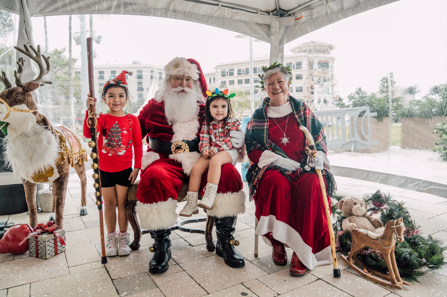 Two young children smiling as they sit with Santa Claus and Mrs. Claus in a festive holiday setup