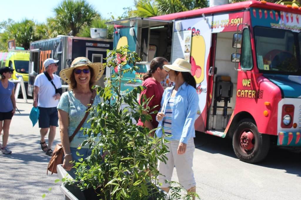 Two women wearing hats and sunglasses are chatting near a colorful food truck with a vibrant mural of a taco and chili peppers, while a third woman tends to plants in a large container