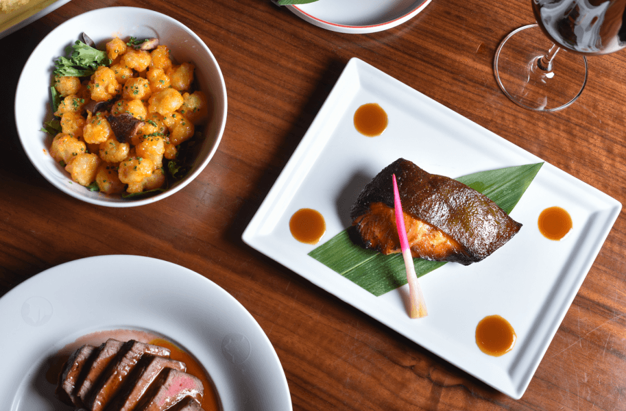 A variety of gourmet dishes including crispy fried balls, grilled fish, and sliced steak with sauces, presented on elegant white plates and bowls on a wooden table