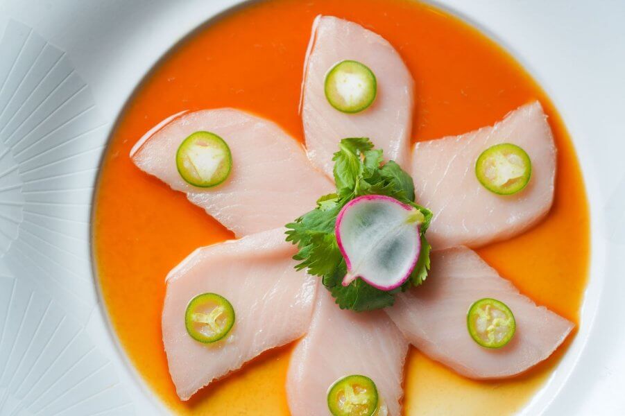 A beautifully presented dish of thinly sliced raw fish arranged in a star pattern on a vibrant orange sauce, garnished with green chili slices and fresh herbs