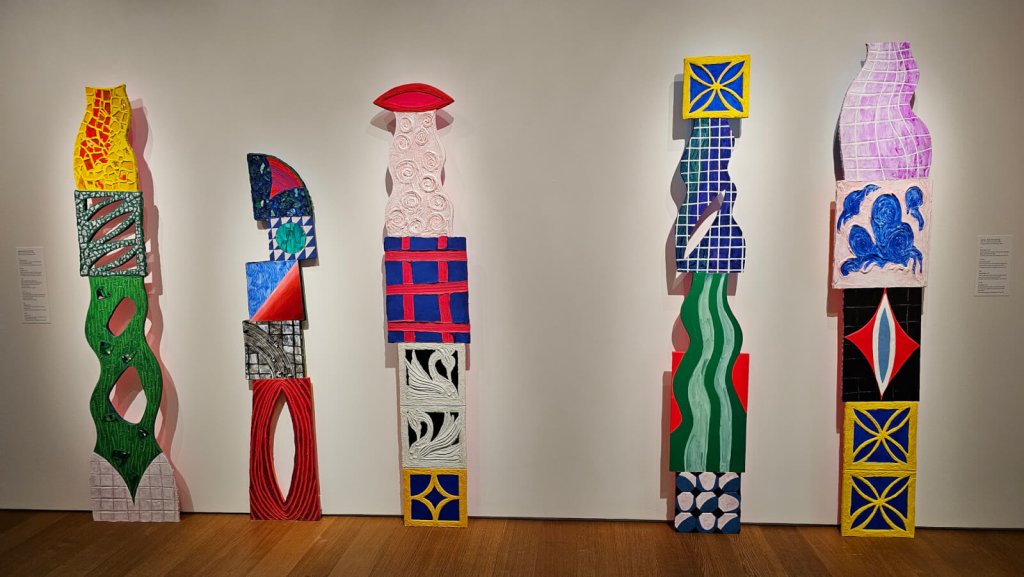 Una serie de cinco esculturas abstractas y llenas de vida, compuestas por formas y motivos geométricos de colores vivos, que se alzan sobre una pared blanca