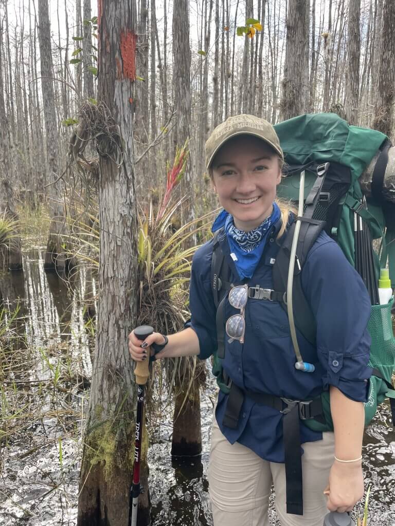 A woman smiles while standing in a swamp, holding a walking stick and wearing a backpack, surrounded by trees and water