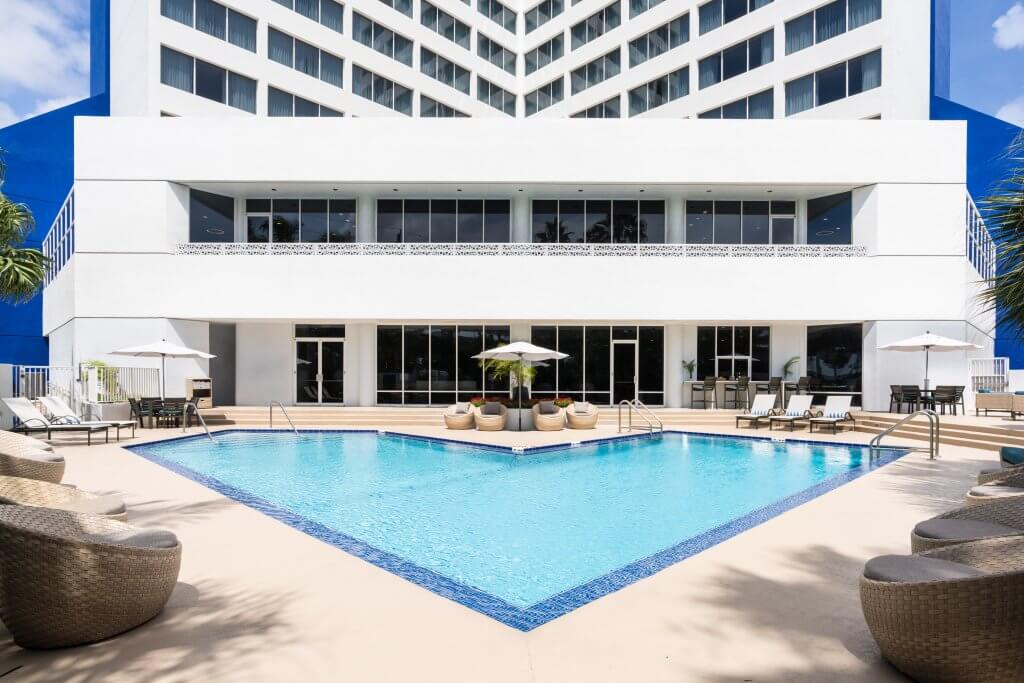 Une luxueuse piscine extérieure entourée de confortables chaises longues et de parasols dans un hôtel moderne à la façade blanche et bleue saisissante.