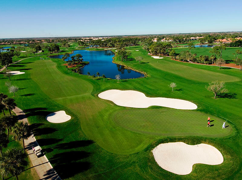 Vista aérea de un frondoso campo de golf con calles sinuosas, búnkers estratégicamente situados y un tranquilo estanque en el que se refleja el cielo azul claro