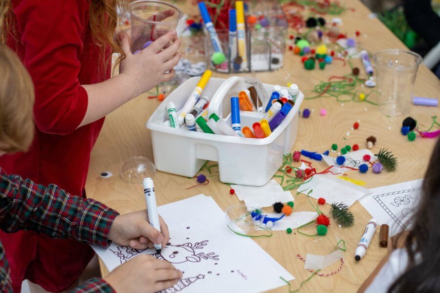 Children engaged in a festive arts and crafts activity, decorating reindeer drawings with colorful markers, pom-poms, and glitter glue