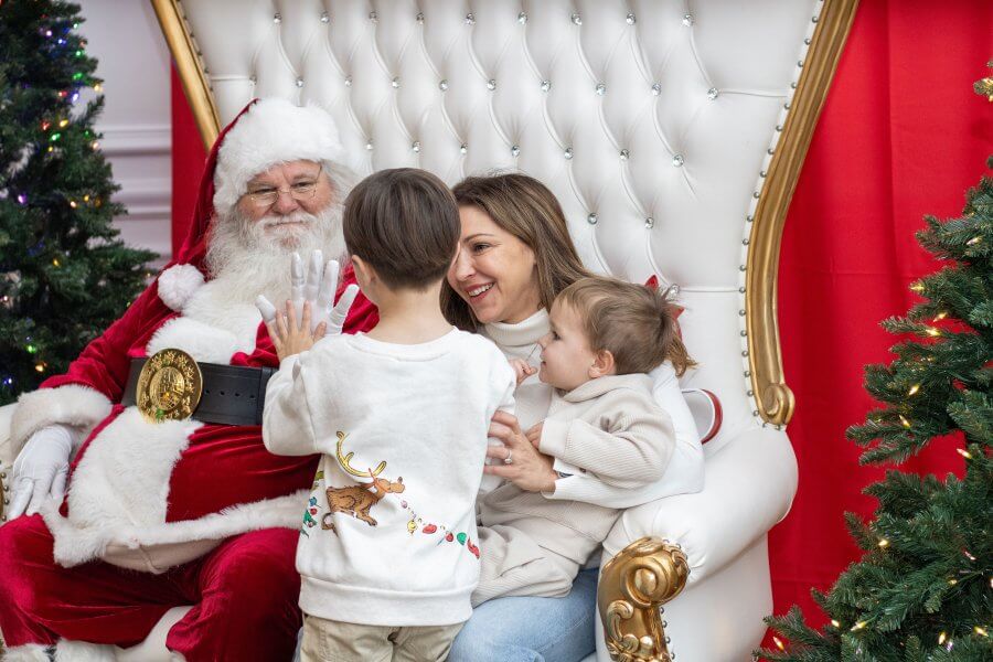 A family of three children and their mother visit Santa Claus in his festive red and white outfit, seated in a golden throne-like chair, surrounded by Christmas trees and decorations