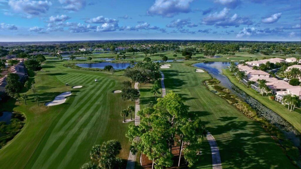 An aerial view of a sprawling golf course with lush green fairways, sand bunkers, and a network of water hazards, surrounded by residential homes and a clear blue sky