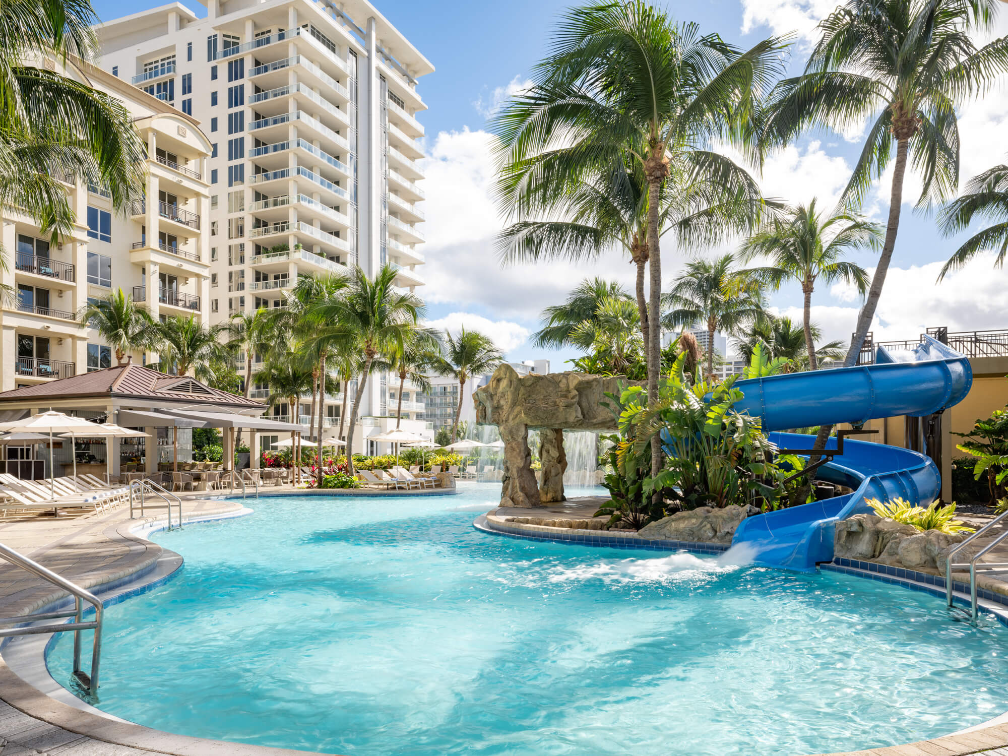 Ein lebhaftes tropisches Resort mit einem großen, einladenden Swimmingpool mit einer blauen Wasserrutsche und einem felsigen Wasserfall, umgeben von üppigen Palmen und modernen Hochhäusern