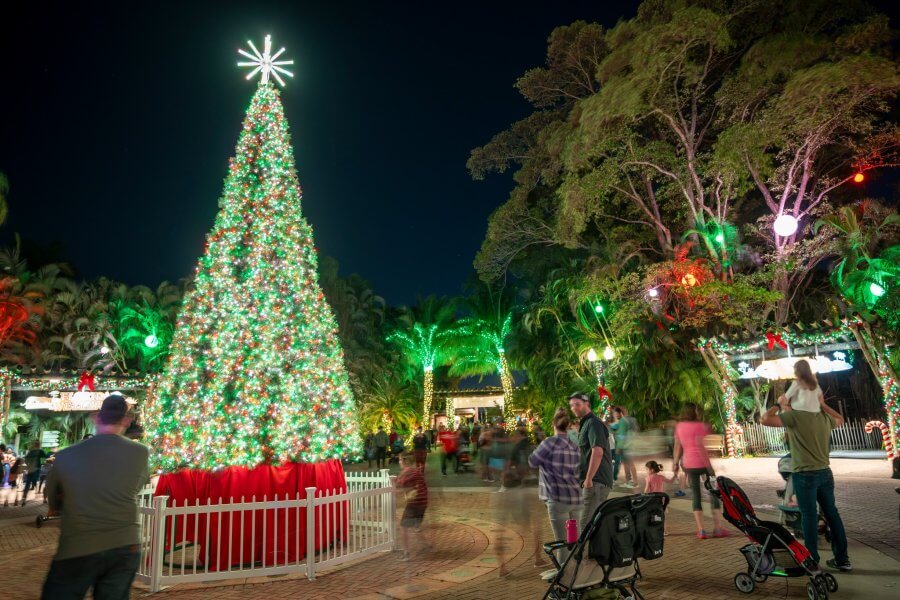 A festive Christmas tree illuminated with vibrant green lights stands tall in a park, surrounded by joyful families and twinkling holiday decorations
