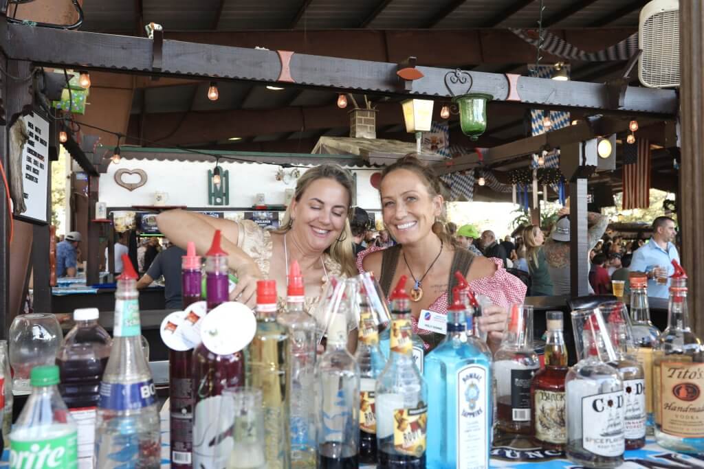 Deux barmaids sourient en versant des boissons dans un bar en plein air décoré de lumières et de drapeaux colorés.