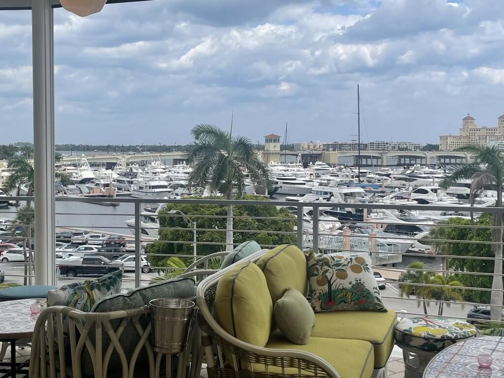 A serene marina filled with numerous white yachts and boats, viewed from a balcony with comfortable seating and a stunning backdrop of palm trees, a bridge, and a historic building