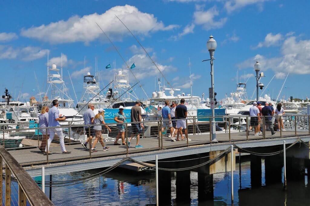 A group of people stroll along a wooden dock lined with luxury boats and yachts in a bustling marina under a clear blue sky