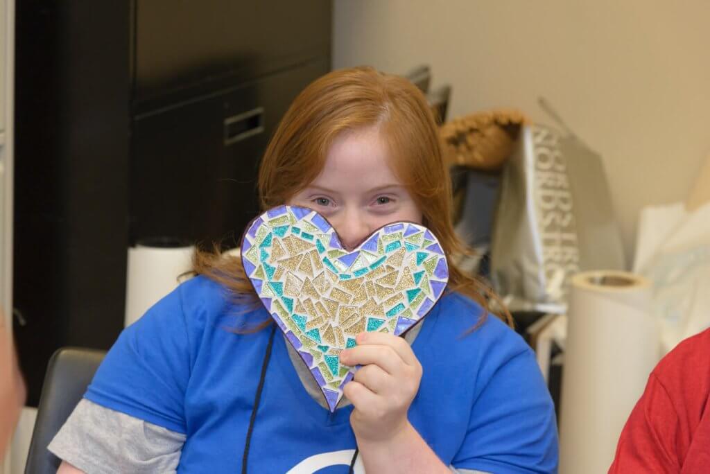 A young girl with red hair is smiling broadly while holding a colorful mosaic heart in front of her face