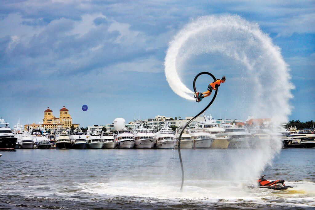 A person performs an aerial stunt on a jetpack over a body of water, creating a spectacular water arc, with a backdrop of luxury yachts and a coastal city skyline