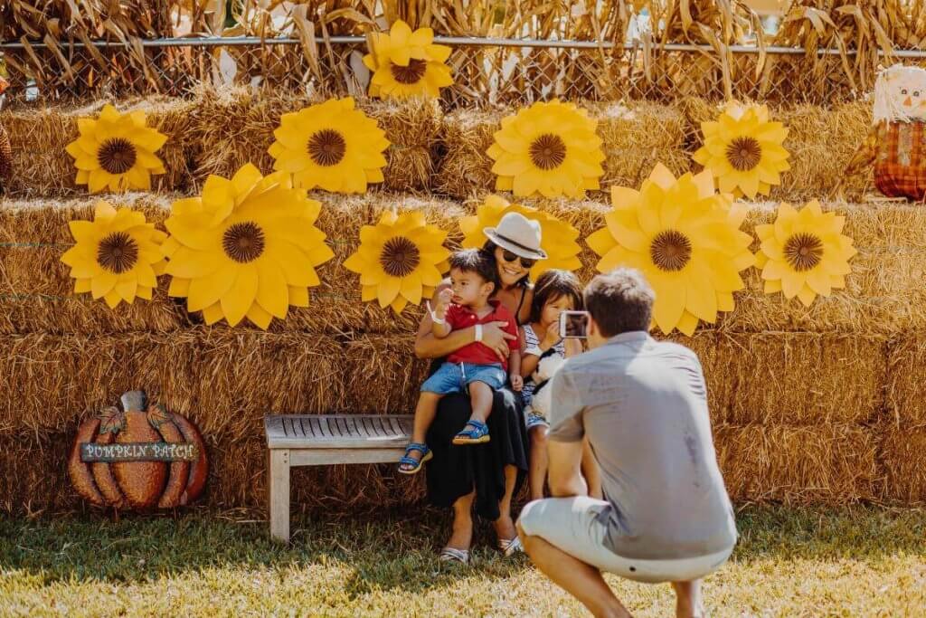 Une famille profite d'une journée ensoleillée dans un champ de citrouilles, posant pour une photo devant une toile de fond de tournesols et de bottes de foin.