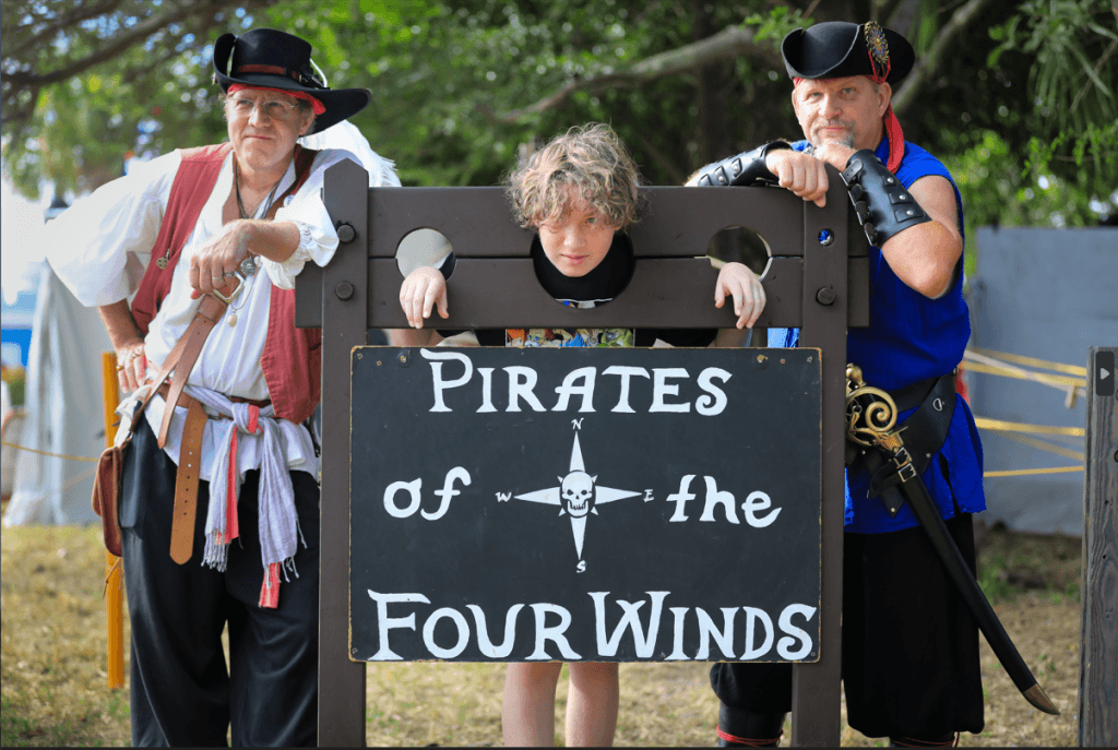 Three individuals dressed as pirates stand behind a sign that reads 'Pirates of the Four Winds', with a young boy peeking through the holes of the sign's wooden frame