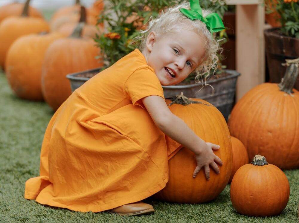 A young blonde girl in an orange dress hugging a large pumpkin