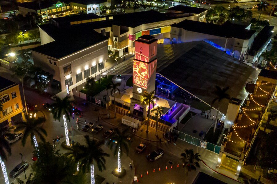 A vibrant nighttime scene of a city square with illuminated buildings, palm trees, and a central clock tower