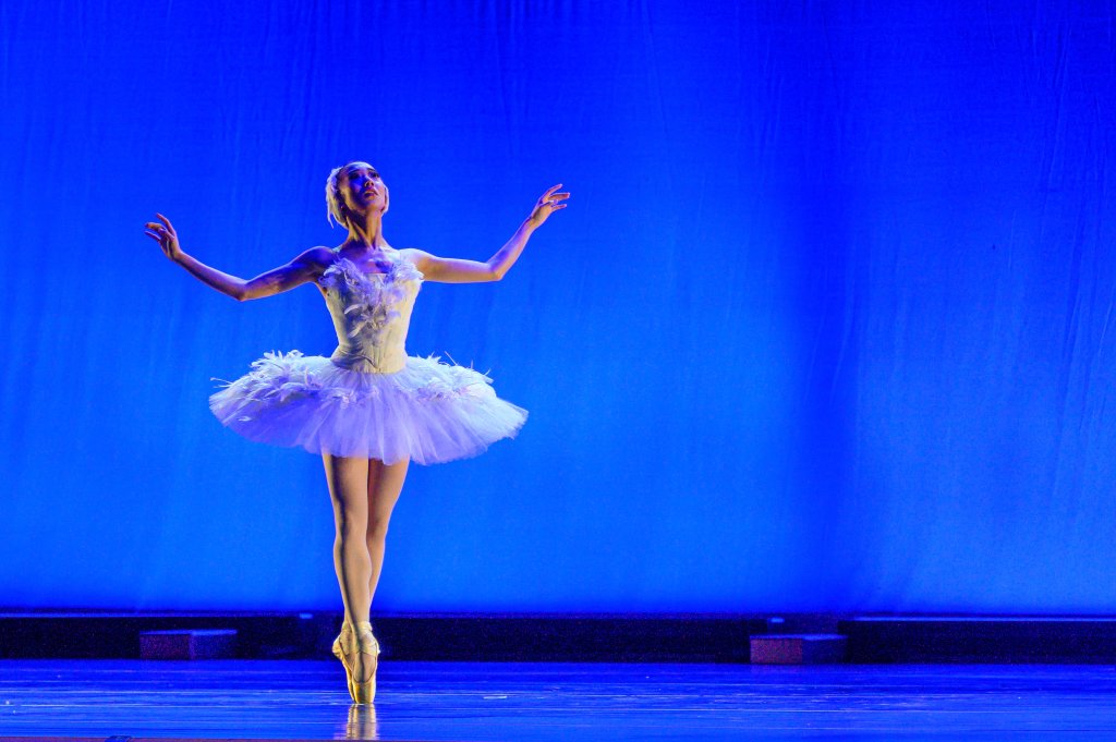 A ballerina in a white tutu performs a graceful leap on stage, illuminated by a vibrant blue spotlight