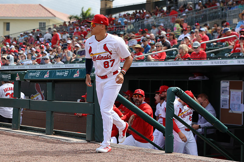 A baseball player wearing a red Cardinals uniform with the number 87 runs on the field, surrounded by a crowd of cheering fans