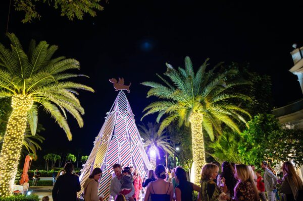 A festive crowd gathers around a colorful, illuminated Christmas tree shaped like a pyramid, surrounded by palm trees and twinkling lights