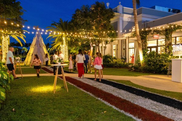 An outdoor evening gathering with people mingling under string lights in a lush garden setting