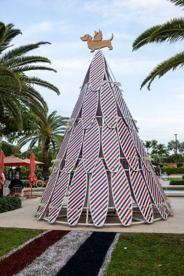 A festive Christmas tree made of surfboards with a dachshund dog topper, surrounded by palm trees and a fountain in a tropical setting