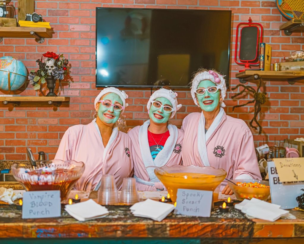 Three ladies wearing a facemask and a pink robe and standing behind a table full of Halloween treats