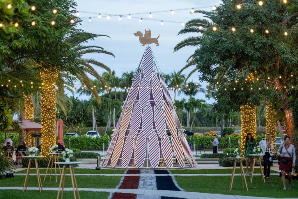 A festive outdoor event with a large, colorful Christmas tree made of surfboards, surrounded by palm trees draped in string lights and guests mingling at high-top tables