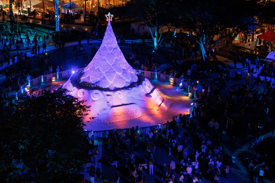 A festive crowd gathers around a massive, illuminated sand sculpture of a Christmas tree in a park at night