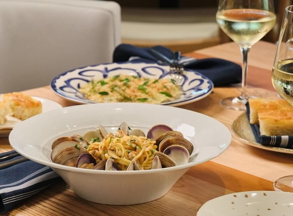 A beautifully set dining table with a variety of Italian dishes, including pasta with clams, risotto, and bread, accompanied by glasses of white wine