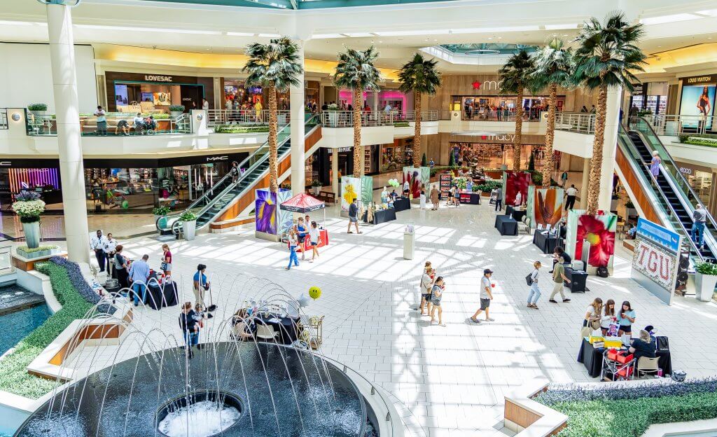 Ein belebtes überdachtes Einkaufszentrum mit mehreren Ebenen, einem großen zentralen Brunnen, Palmen und einer Vielzahl von Geschäften und Restaurants