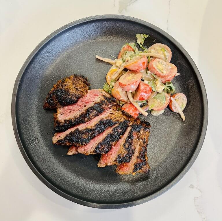 Un jugoso filete a la parrilla con los bordes ligeramente chamuscados, acompañado de una fresca ensalada de tomate y hierbas, servido en un elegante plato negro