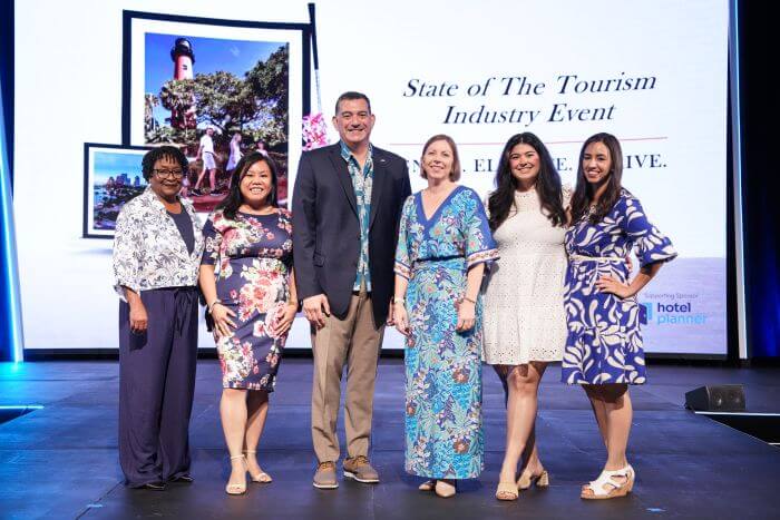 Six individuals pose together on a stage at a tourism industry event