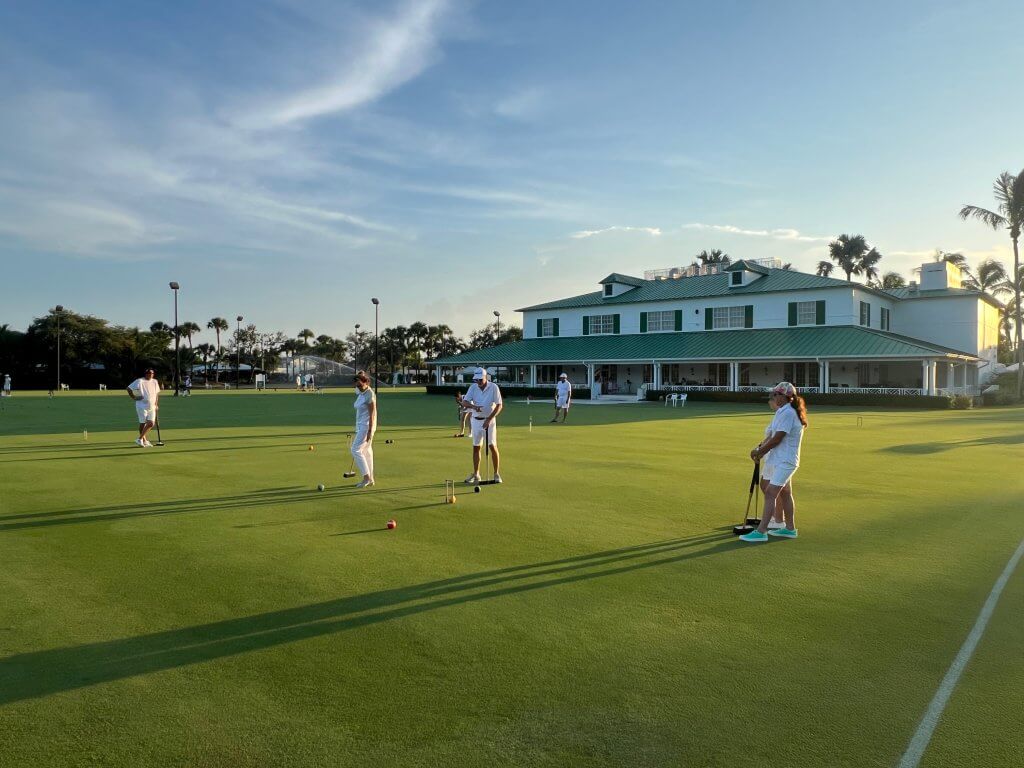 A group of people dressed in white are playing croquet on a lush green lawn in front of a large white house with a green roof