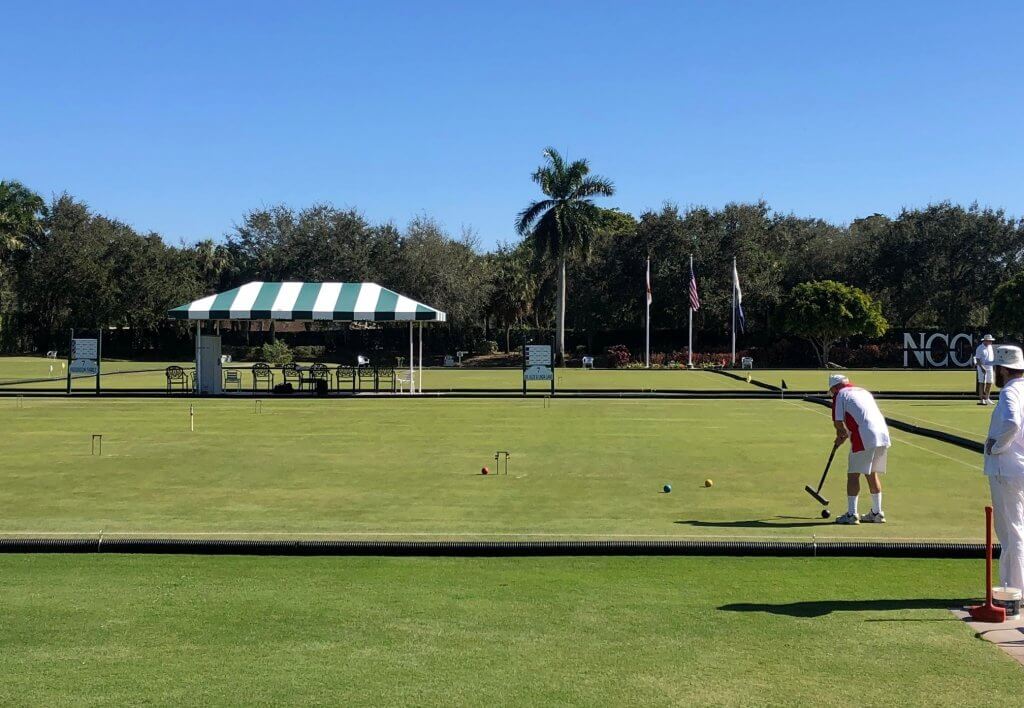 A player in white attire is carefully aiming a croquet mallet at a ball on a well-manicured green court, with spectators and flags in the background
