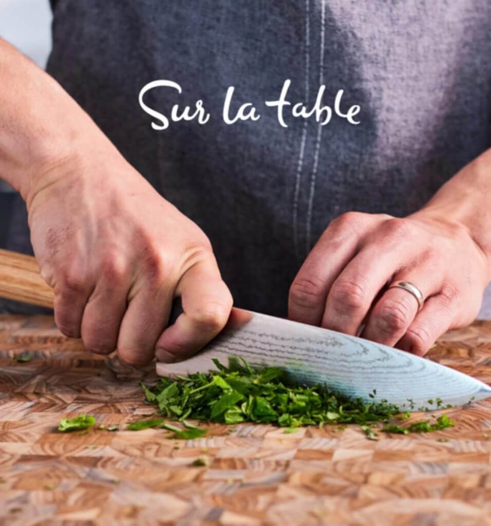 A person wearing a 'Sur la table' apron finely chops fresh herbs with a sharp knife on a wooden cutting board