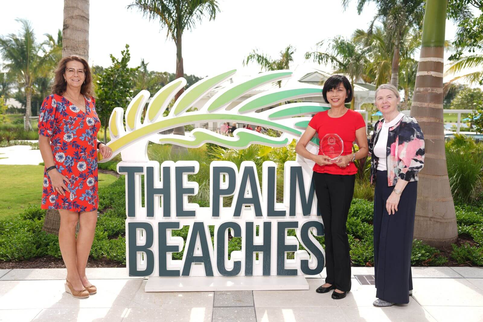 Three women stand in front of a large sign for The Palm Beaches, with one holding an award