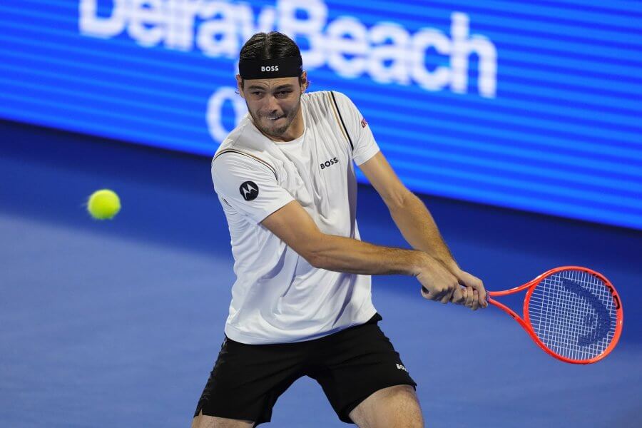 A tennis player in a white shirt and black shorts is preparing to hit a tennis ball with his racket on a blue court
