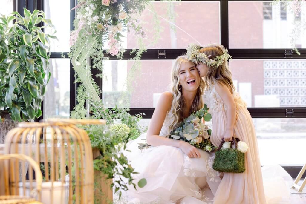 A joyful bride and flower girl share a tender moment amidst a lush, green backdrop with cascading flowers and natural light streaming through large windows