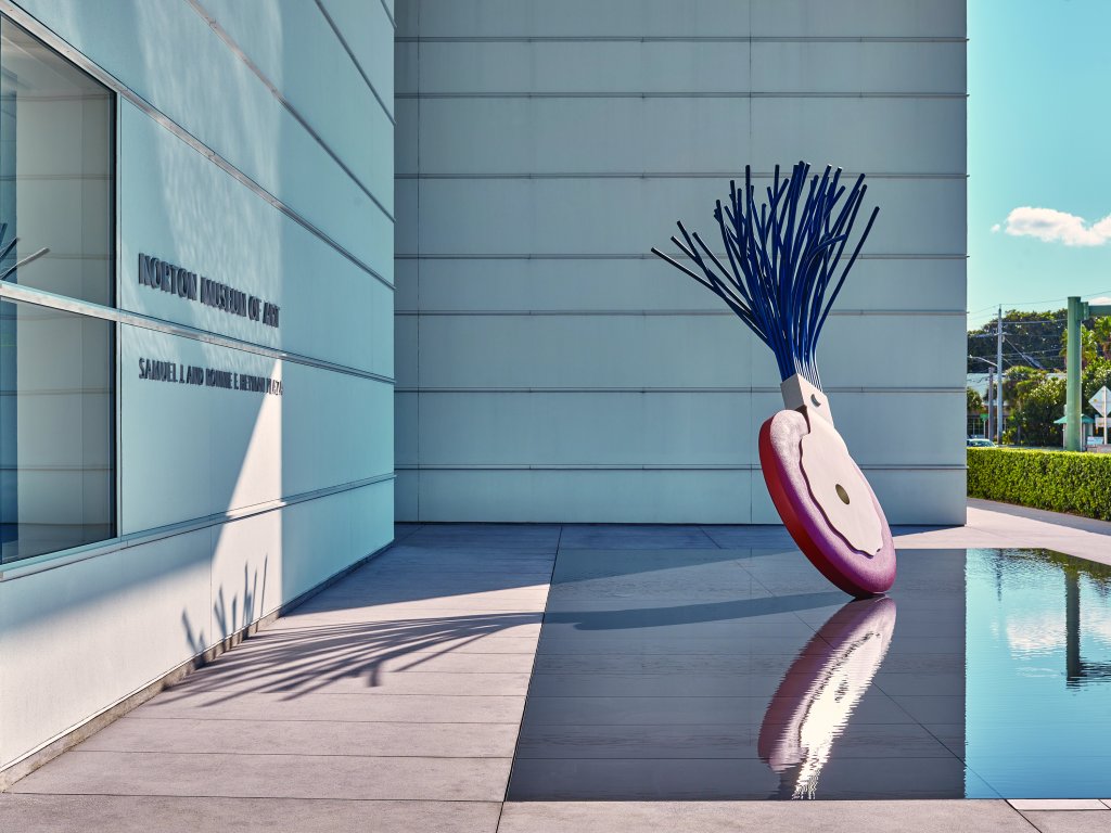 Una gran escultura abstracta con una base roja en forma de corazón y protuberancias azules en forma de púas se erige frente a un edificio moderno, con un estanque reflectante cerca