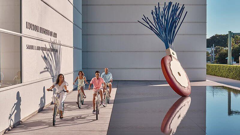 A family of four enjoys a leisurely bike ride along a sunny path beside a modern art museum, with a striking blue and white sculpture reflected in a nearby pool