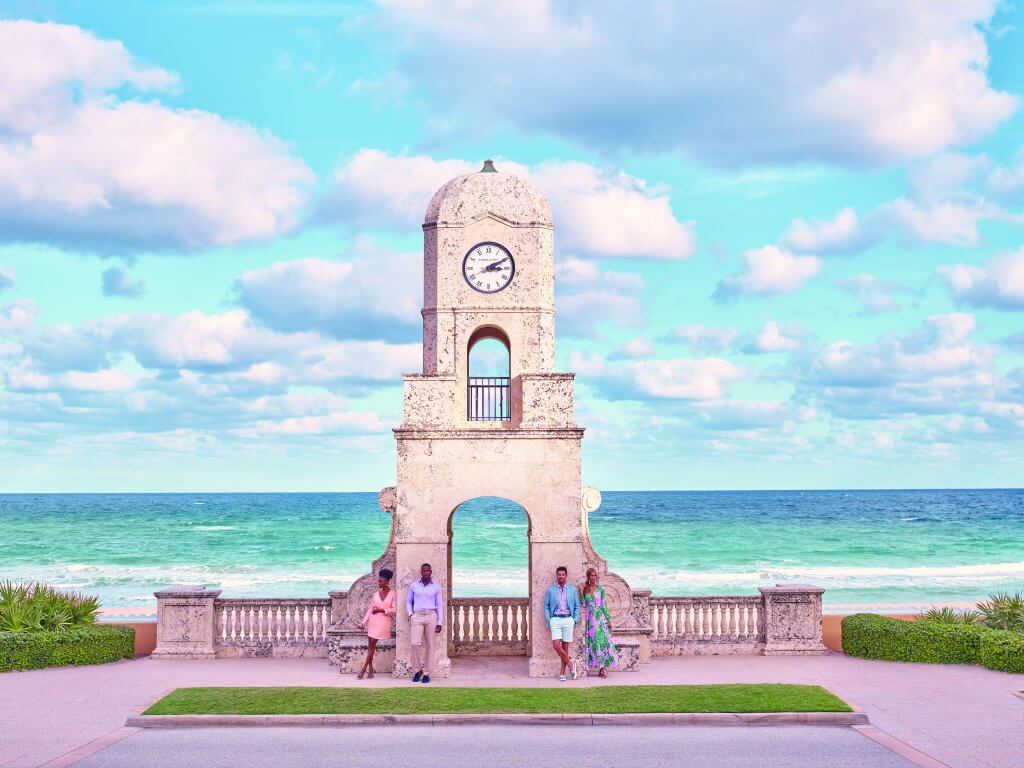 Une famille de quatre personnes se tient devant une pittoresque tour d'horloge surplombant l'océan, avec un ciel d'un bleu éclatant et des nuages cotonneux au-dessus.