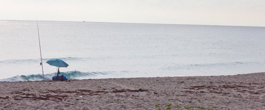 Una tranquila escena de playa con una única sombrilla azul y una caña de pescar en la arena, con vistas a un mar en calma