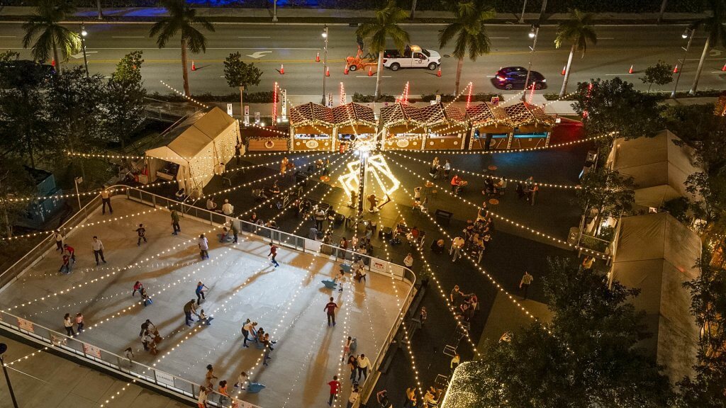 A festive outdoor ice rink illuminated by twinkling lights, surrounded by palm trees and holiday market stalls