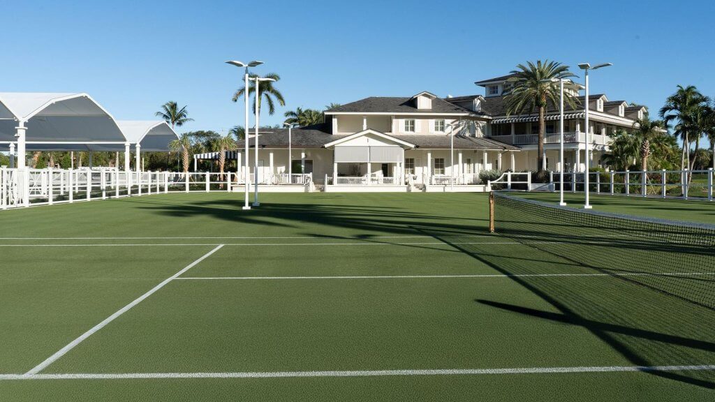 Un court de tennis serein avec une surface verdoyante, des lignes blanches et un filet, avec en toile de fond un grand manoir blanc avec un porche et des palmiers.