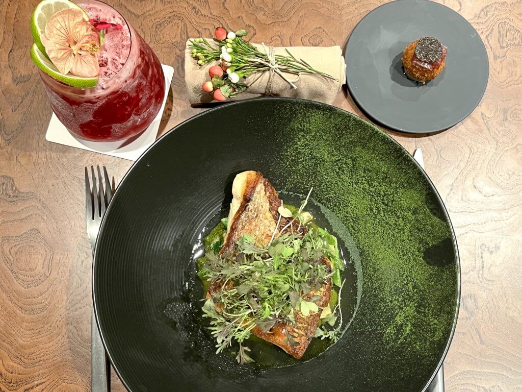 Un plato con una presentación exquisita que incluye un trozo de pescado adornado con microverduras y servido sobre una base de puré verde, acompañado de un cóctel de un rojo intenso con adornos de lima y flores