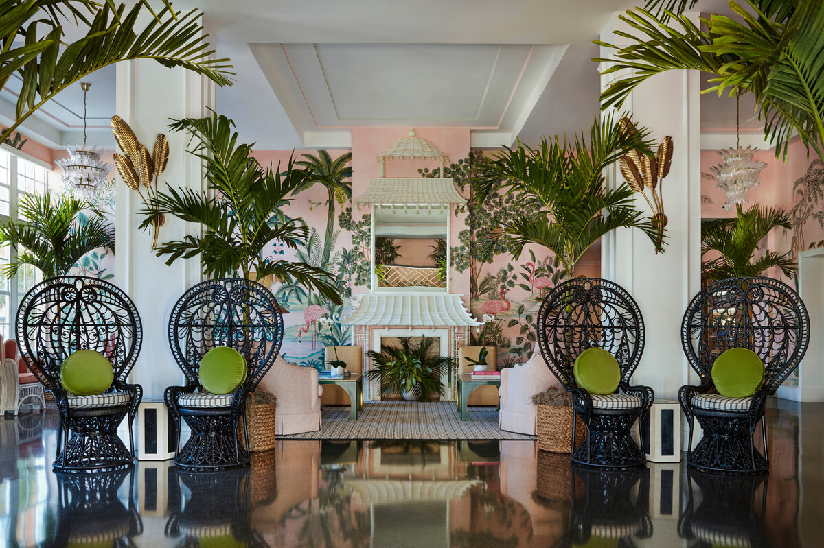 Un hall d'entrée luxueux au thème tropical, avec des chaises en osier noir, des coussins verts et une peinture murale rose.