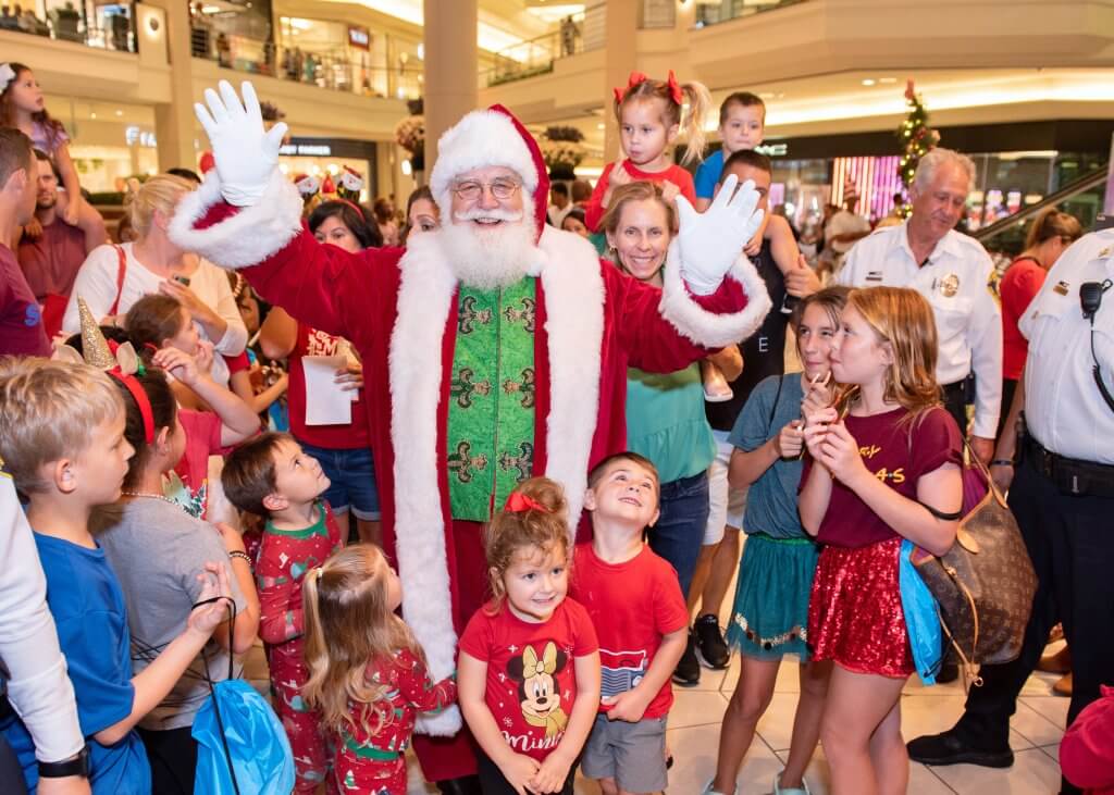 Santa Claus waves to a crowd of excited children and families in a bustling mall, surrounded by festive decorations and holiday cheer